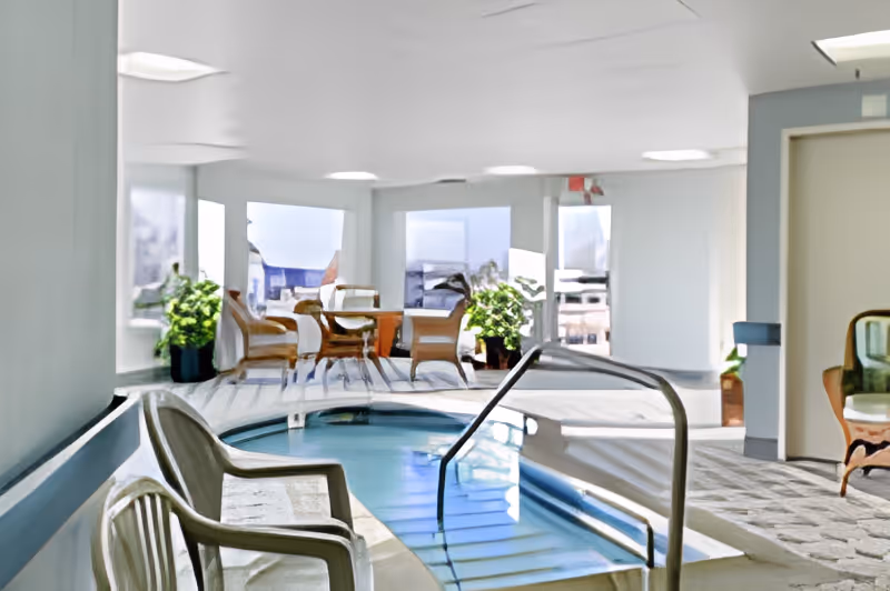 Indoor pool area with a small rectangular pool featuring handrails and steps leading into the water. Surrounding the pool are several chairs and tables, with large windows allowing natural light to fill the space. Potted plants add greenery to the bright, clean room.