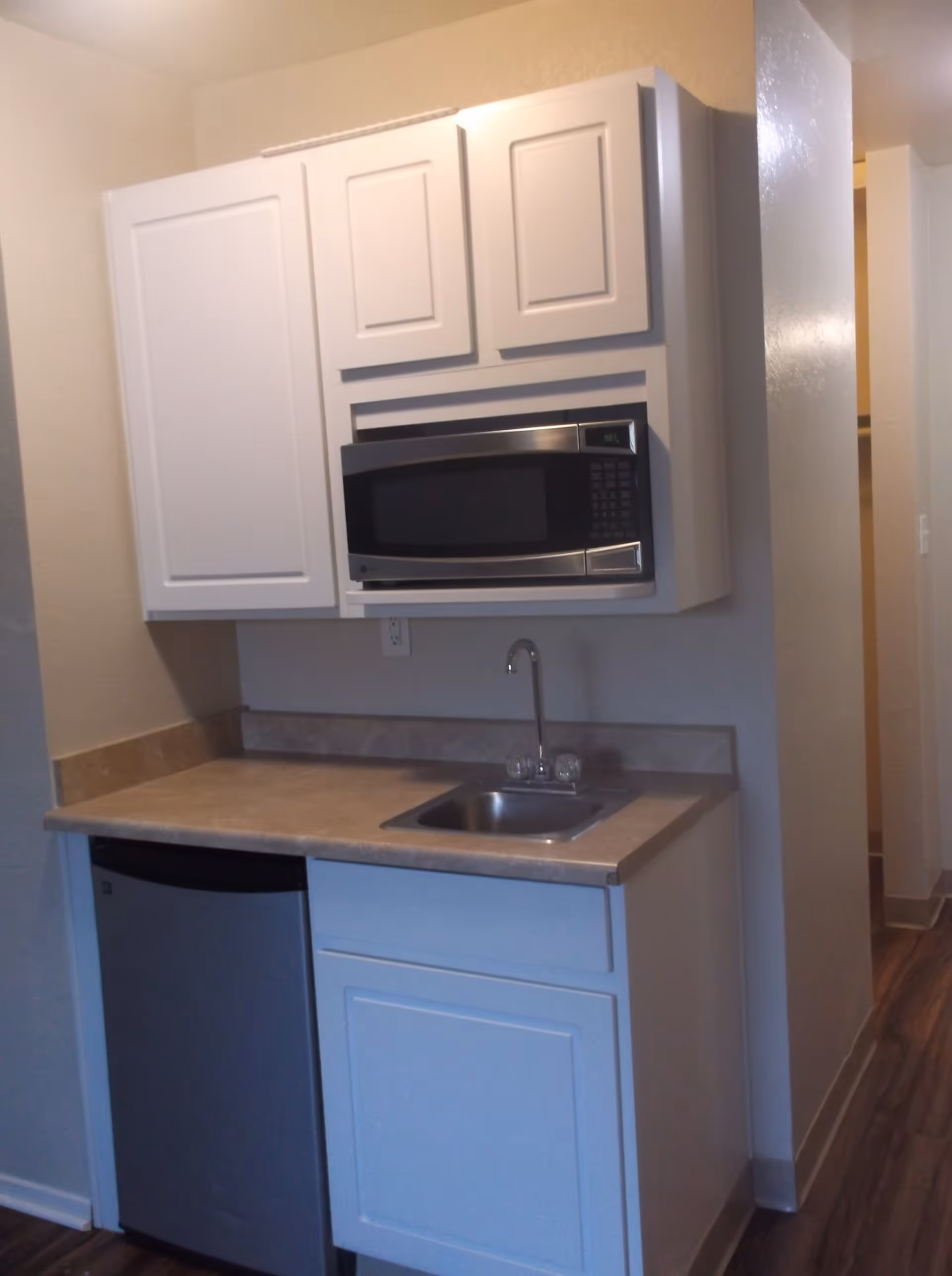 Small kitchenette with white cabinets, a stainless steel microwave mounted above the countertop, a small sink with a faucet, and a compact refrigerator below the counter. The floor is wood-style laminate and the walls are painted light beige.