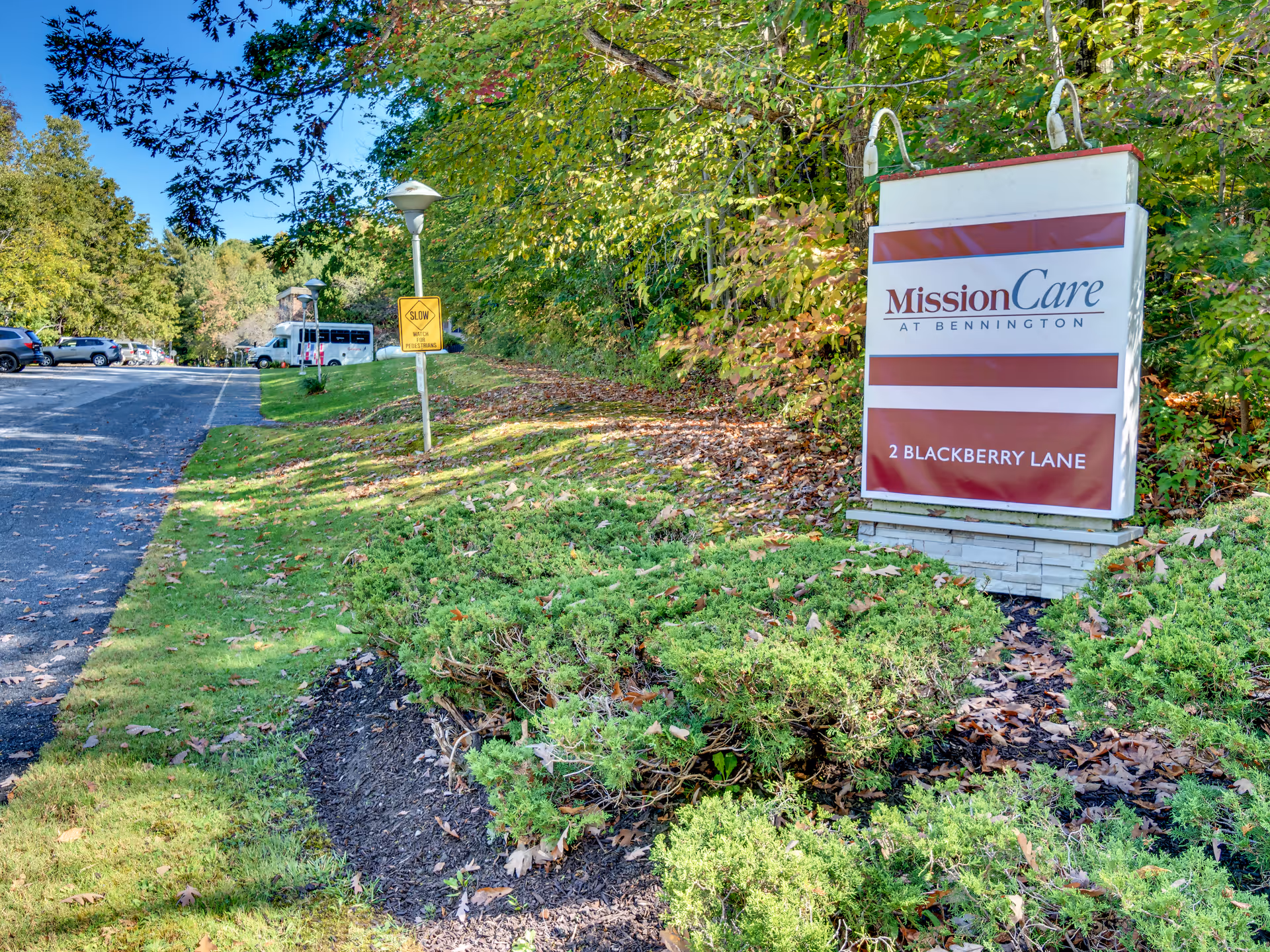 Outdoor view of the entrance area to MissionCare at Bennington featuring a large sign with the facility name and address 2 Blackberry Lane, surrounded by green bushes and trees with a parking lot and vehicles visible in the background.