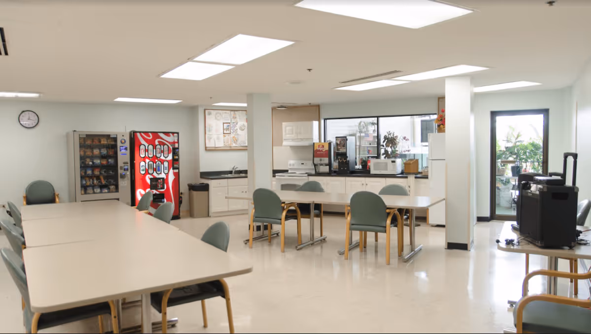 A bright communal room with tables and chairs arranged for group seating, vending machines against the wall, a kitchenette area with a stove, microwave, coffee machine, and cabinets, and a door leading to an outdoor space with plants.