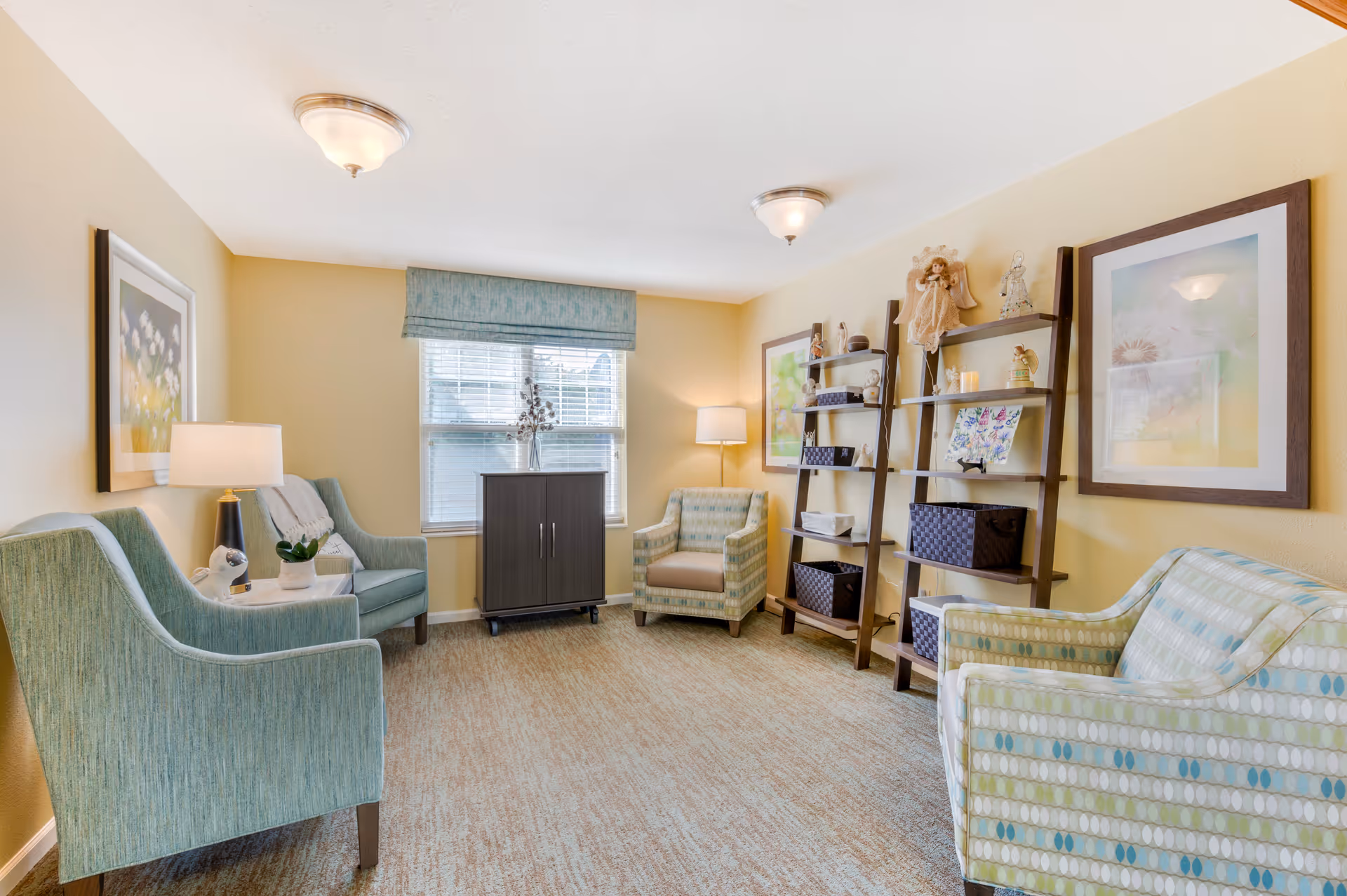 Sunlit lounge with patterned upholstered chairs arranged around a window, decorative shelving, framed artwork, and soft lighting.