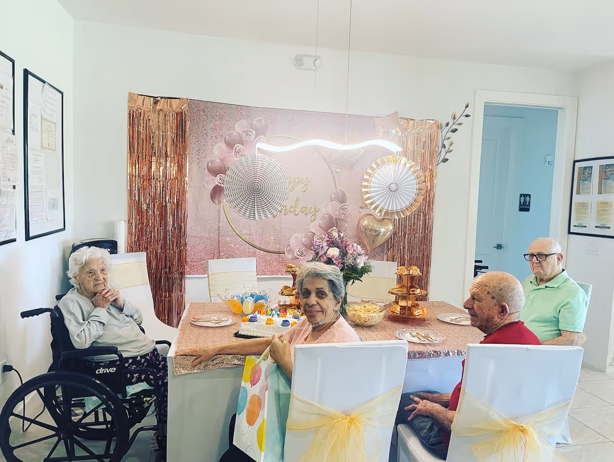 Four elderly people sitting around a decorated table in a room. The table is covered with a pink tablecloth and has party decorations including a cake, flowers, and balloons. One person is in a wheelchair, and the others are seated on white chairs with yellow bows. The background features a pink and gold birthday backdrop with balloons and paper fans.