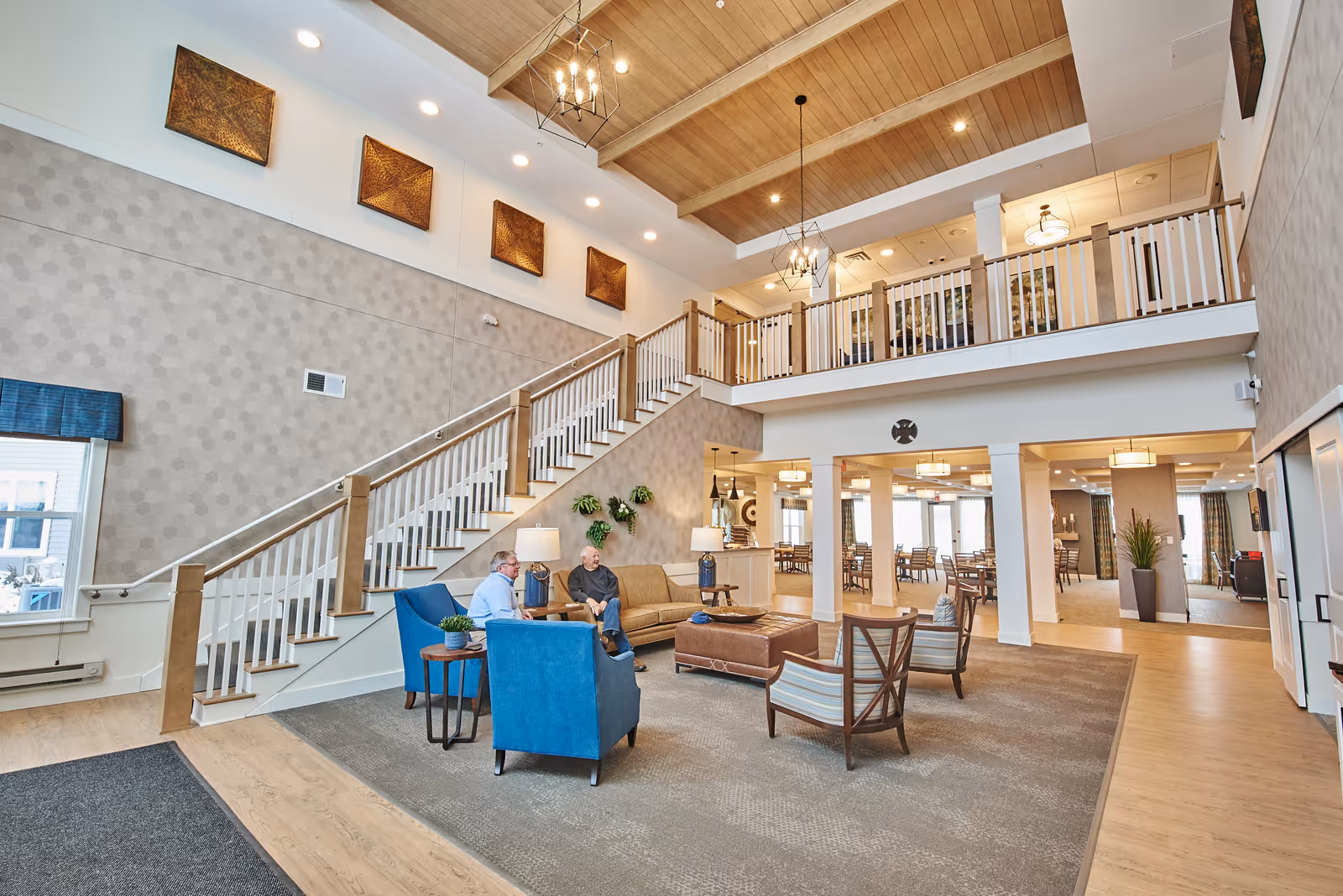 Bright two-story communal lounge with seating, a staircase, and a dining area in a senior living facility.