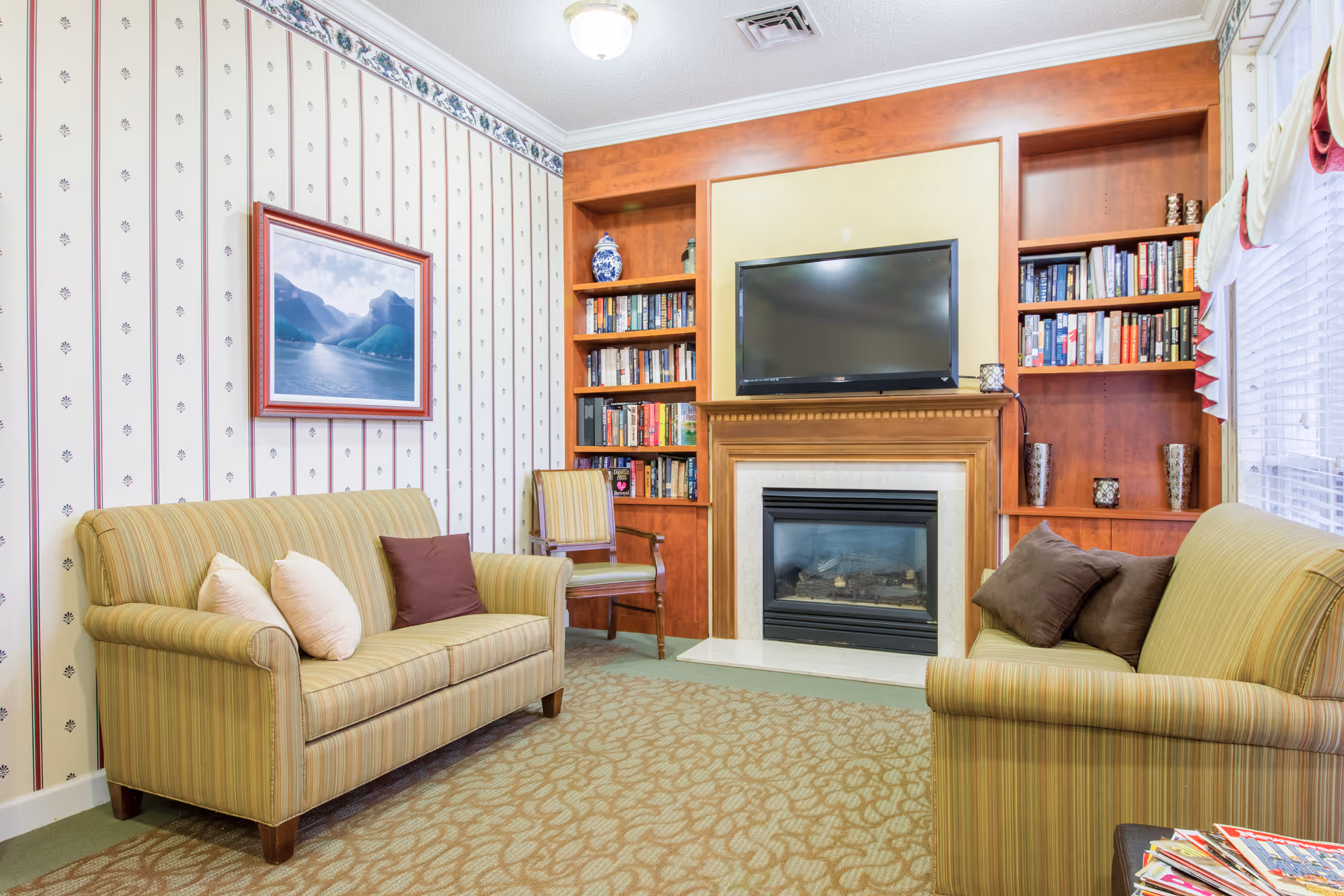 A cozy sitting room with two striped sofas facing a fireplace and wall-mounted TV flanked by built-in bookshelves.
