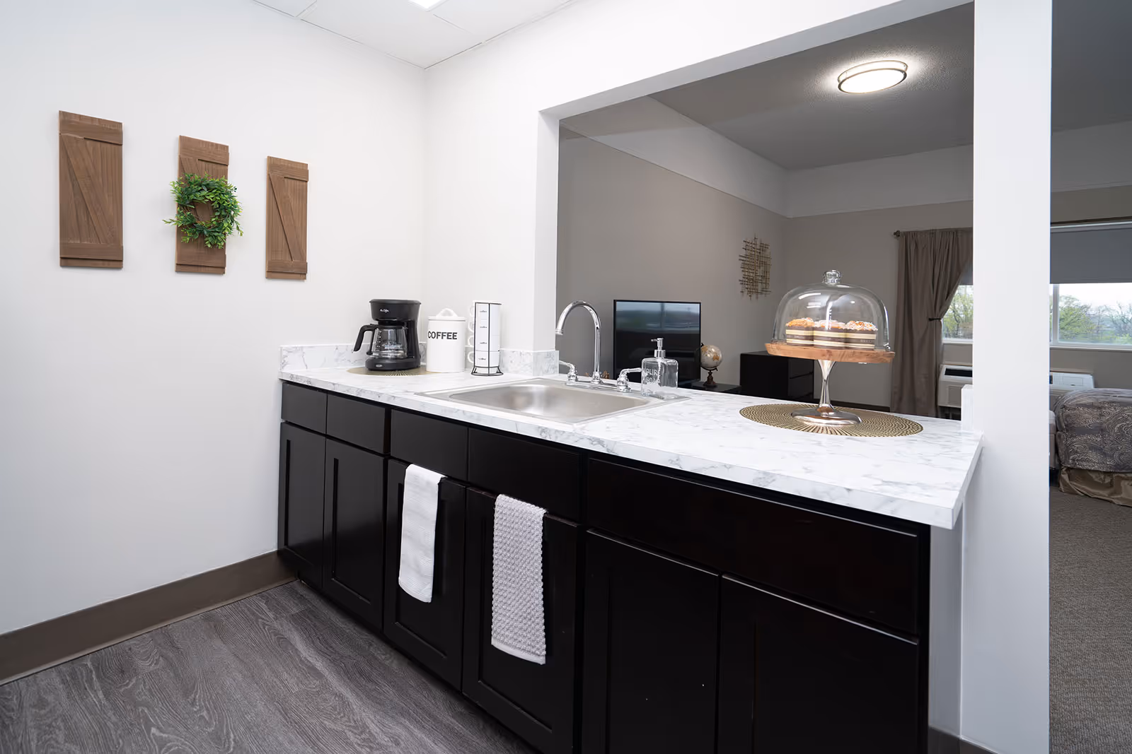 Small kitchenette with a marble countertop, sink, coffee maker, dark cabinets and a passthrough to a living area.