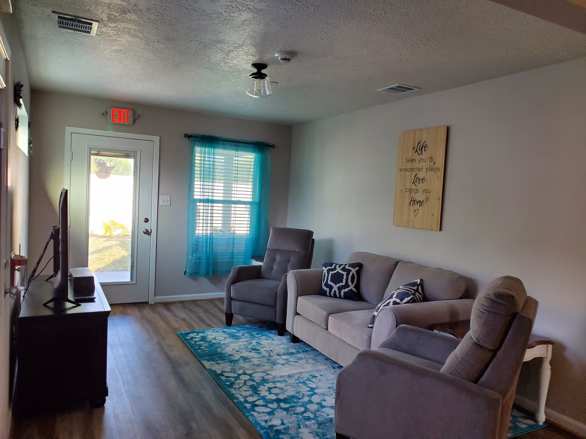 A cozy living room with a gray couch and two matching armchairs arranged around a blue patterned rug. A wooden TV stand with a flat-screen TV is positioned opposite the seating. A window with sheer turquoise curtains lets in natural light. A wooden wall plaque with an inspirational quote hangs above the couch. The room has light-colored walls and wood flooring, with an exit door leading outside.