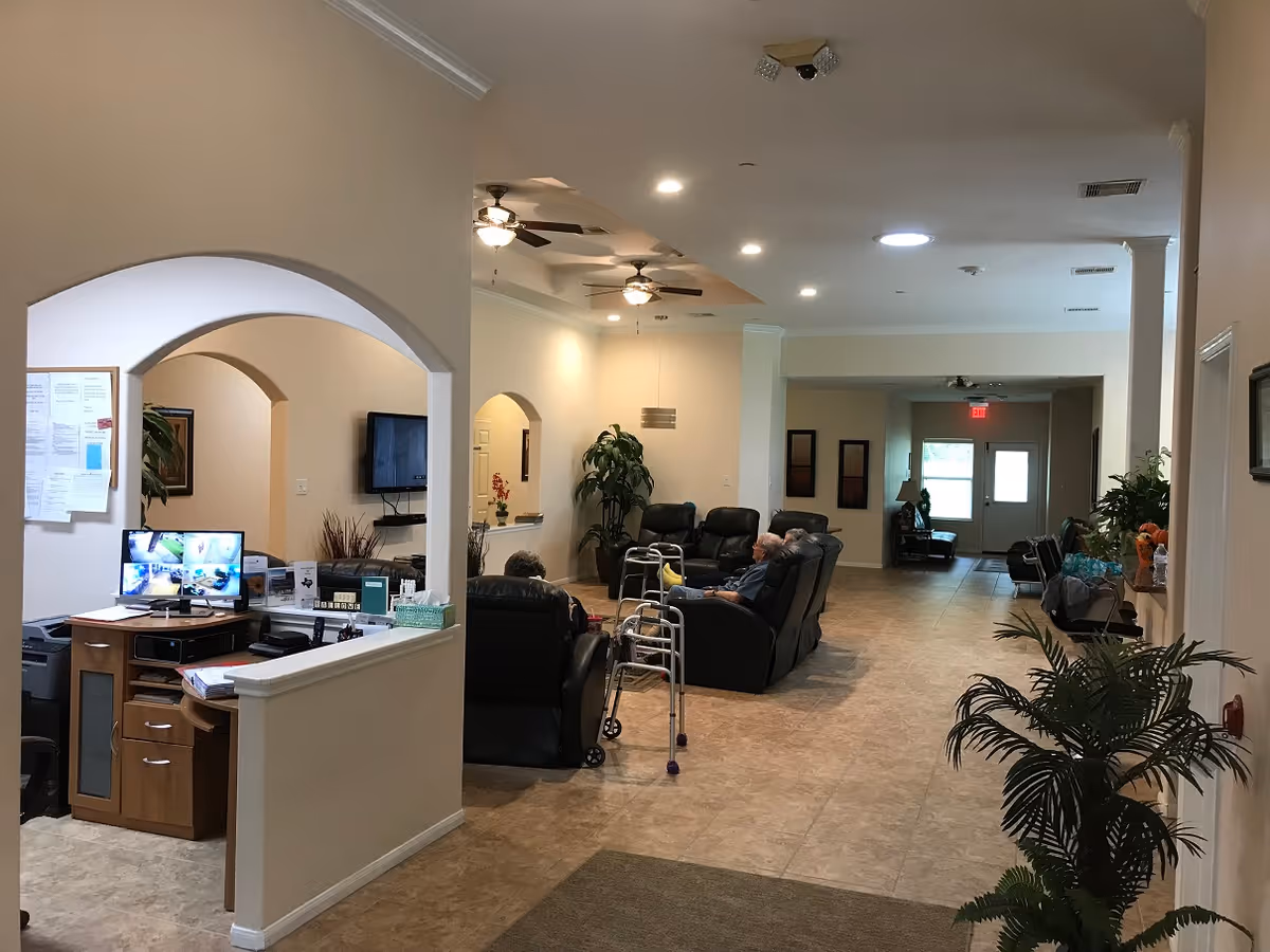 Interior view of a senior living facility lounge area with several elderly people seated in black recliners. The room has beige walls, tiled floors, ceiling fans, and recessed lighting. There is a reception desk with computer monitors on the left side and various plants and decorations throughout the space.