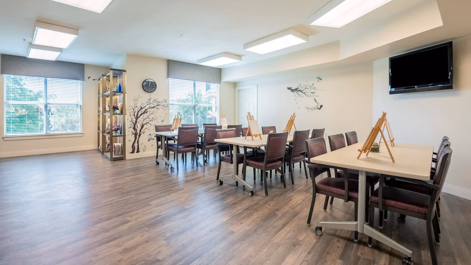 A bright and spacious activity room with multiple tables and chairs arranged for group activities. Each table has small wooden easels, suggesting it is set up for painting or drawing. The room has large windows with blinds, a wall clock, decorative wall art of trees and birds, a shelving unit with decorative items, and a wall-mounted flat-screen TV.