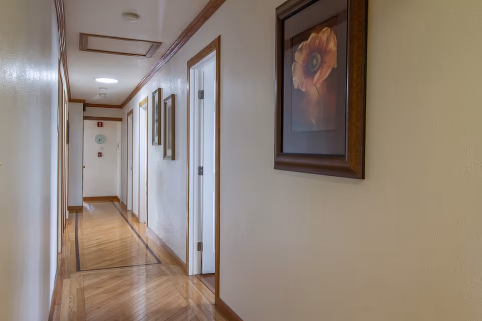 A well-lit hallway with polished wooden floors and cream-colored walls. Several doors line the right side of the hallway, and framed floral artwork is hung on the walls. The ceiling has wooden trim and recessed lighting.