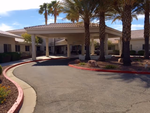 Entrance driveway with a covered drop-off area at a single-story building surrounded by palm trees and desert landscaping under a blue sky.