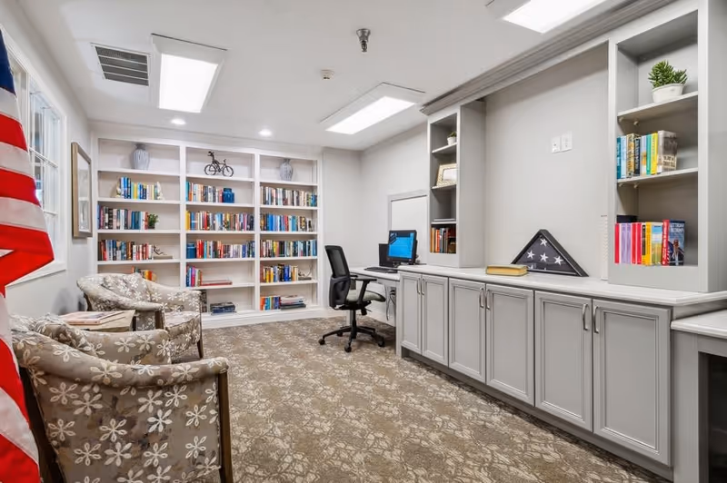 A cozy interior room with built-in bookshelves filled with books, two floral-patterned armchairs, a desk with a computer and office chair, and cabinets with decorative items including a folded American flag in a display case. The room has a carpeted floor and bright overhead lighting.