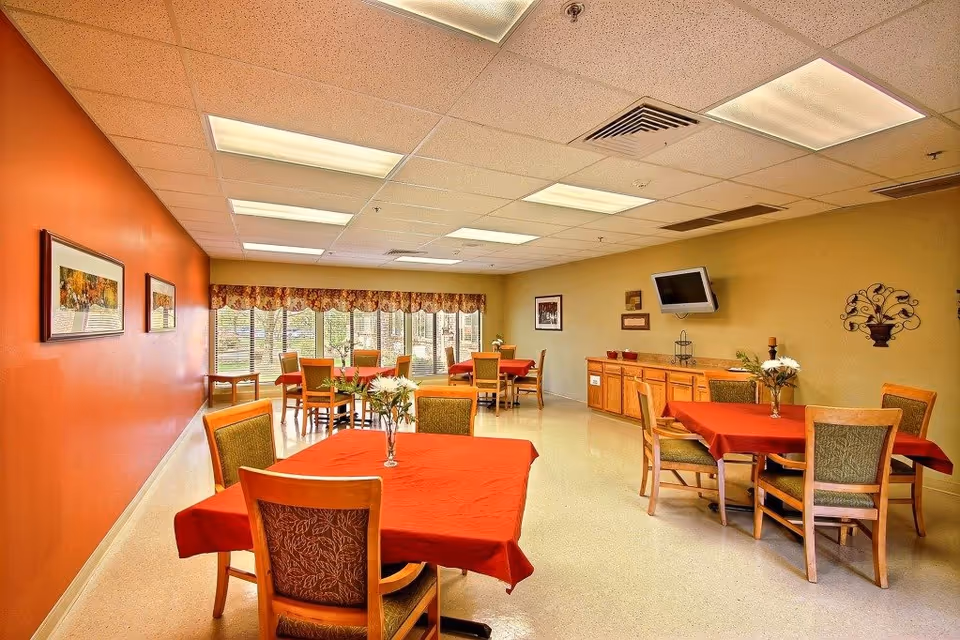 A dining room in Greenwood Healthcare Center with several tables covered in red tablecloths and wooden chairs with green upholstery. The room has an orange accent wall with framed artwork, large windows with floral valances, a wall-mounted TV, and a wooden sideboard with decorative items.