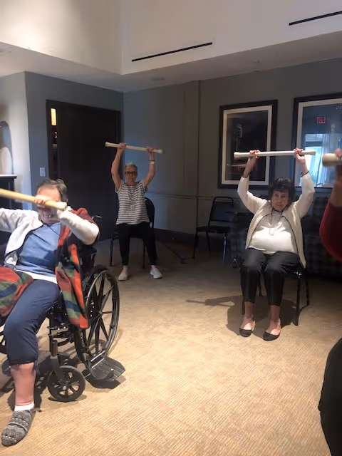 Three elderly women seated indoors participating in a group exercise, each holding a wooden stick above their heads. One woman is in a wheelchair, and the room has beige carpet and neutral-colored walls with framed pictures.
