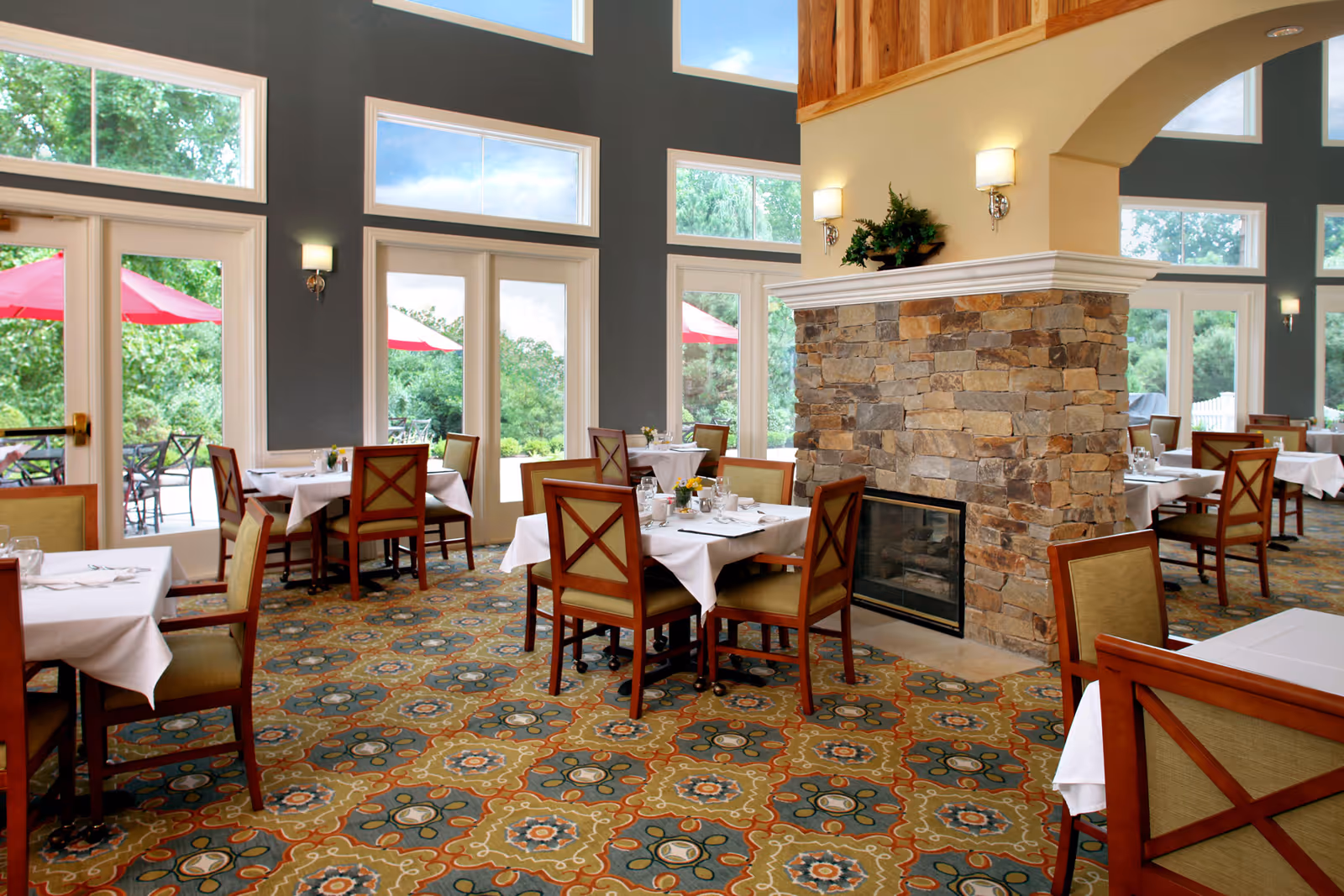 Dining room with tables set with white tablecloths, wooden chairs, patterned carpet, large windows, and a central stone fireplace.