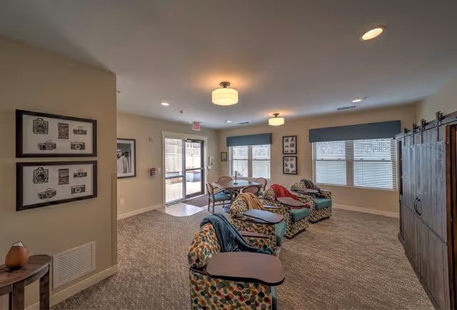 A cozy senior living common area with patterned armchairs equipped with attached wooden trays, a round table with chairs near large windows, and soft overhead lighting. The room has beige walls, carpeted floors, framed artwork on the walls, and a large wooden cabinet on the right side.