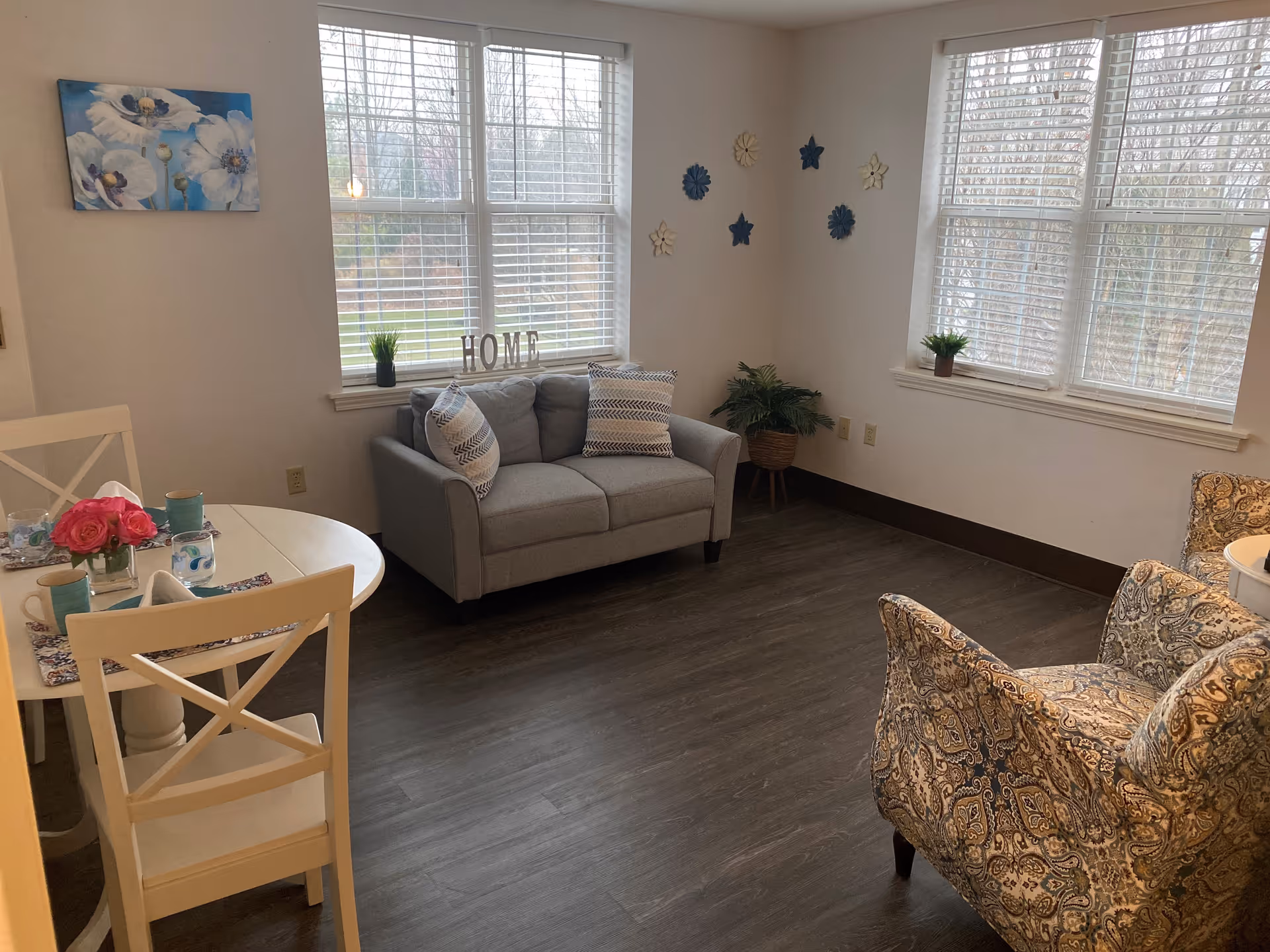 A cozy living room area with a small gray loveseat adorned with two patterned pillows, a patterned armchair, and a white round dining table with two chairs. The table is set with cups, napkins, and a vase of pink flowers. The room has large windows with white blinds letting in natural light, wall decorations including blue and white star-shaped ornaments, a small plant on the windowsill, and a painting of white flowers on the wall.