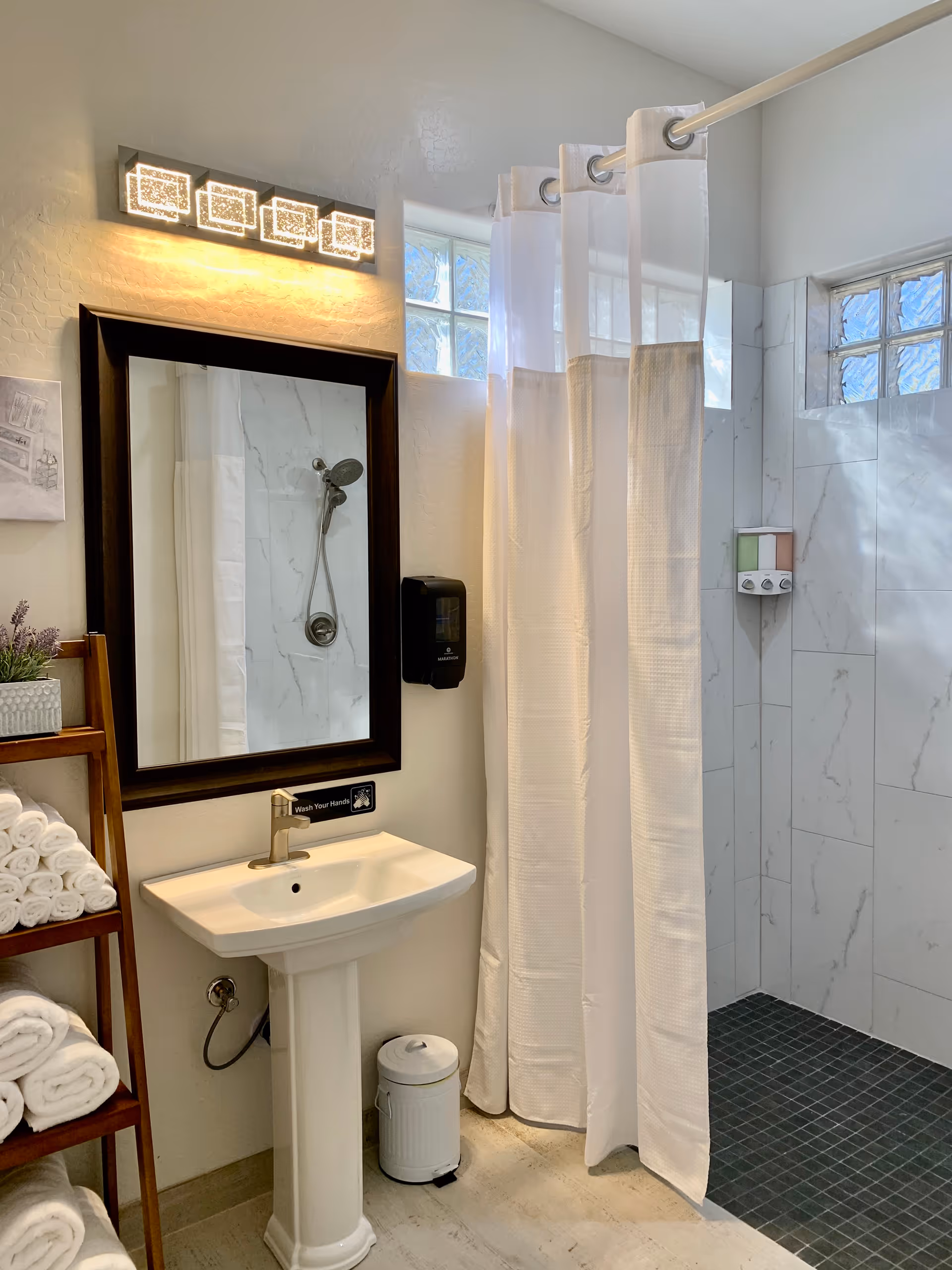 Well-lit bathroom featuring a pedestal sink and mirror, a walk-in tiled shower with a white curtain, and a ladder shelf stocked with rolled towels.