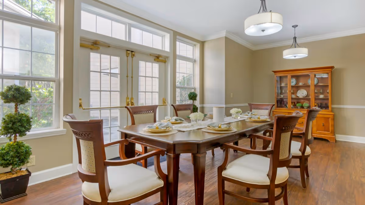 Sunlit dining room with a wooden table set for six, upholstered chairs, a china cabinet, and large windows with double doors.