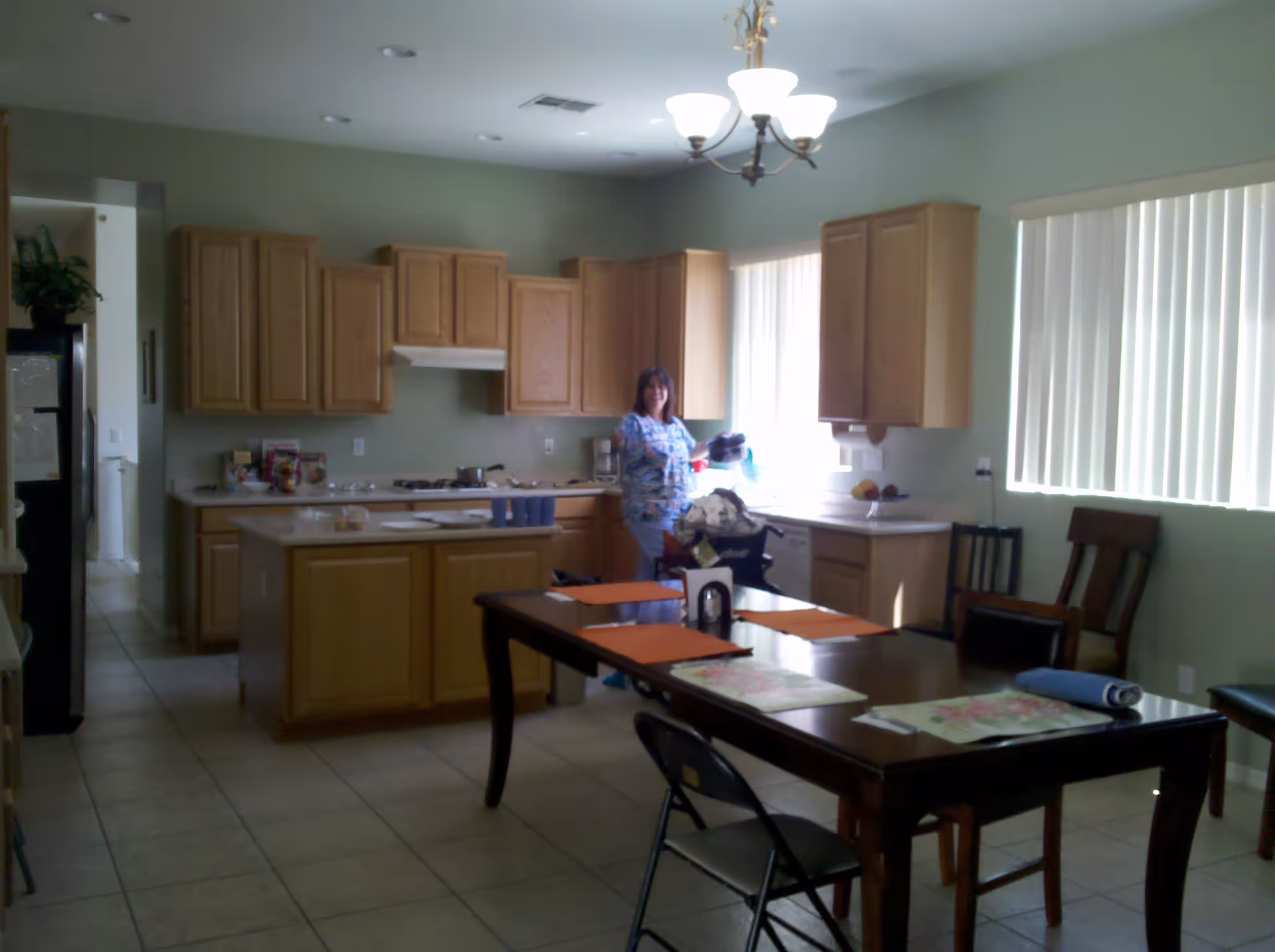 A kitchen and dining area with wooden cabinets, a stove, and a refrigerator. A woman stands near the counter holding an object. The dining table has placemats and chairs around it. Vertical blinds cover the windows, and the room is lit by a ceiling light fixture.