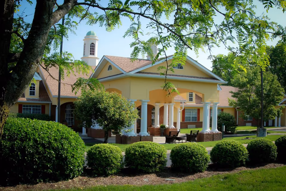 Exterior view of a senior living facility building with a covered entrance supported by white columns, surrounded by green bushes and trees under a clear blue sky.