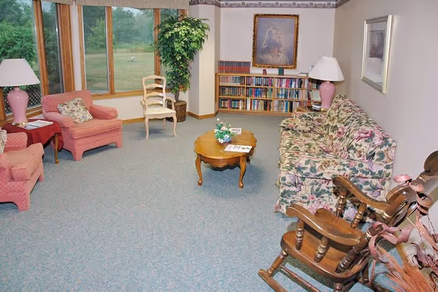 A cozy living room with floral patterned sofa, two pink armchairs, a wooden rocking chair, and a wooden coffee table with a small flower arrangement and magazines. There are two table lamps with pink bases, a bookshelf filled with books, a framed portrait on the wall, and large windows showing a green outdoor view.