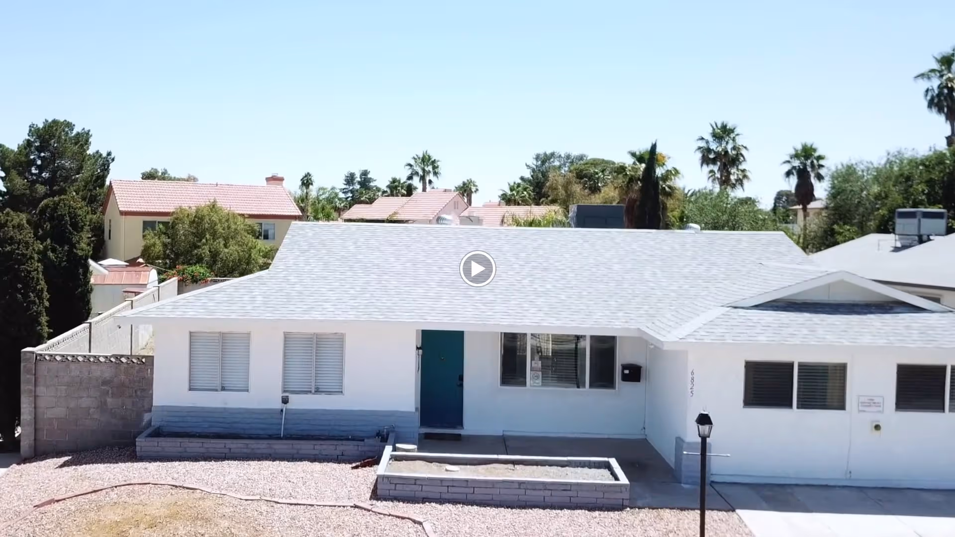 Front exterior view of a single-story white building with a gray shingled roof, a teal front door, and several windows with shutters. There is a small raised garden bed and a lamp post in front of the building. Trees and other houses are visible in the background under a clear blue sky.