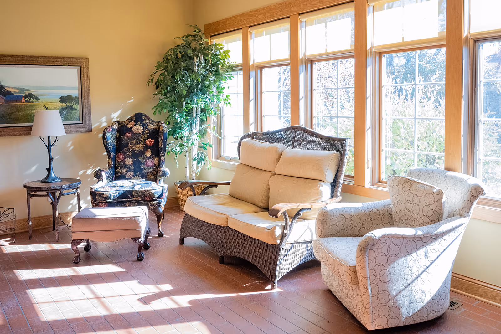 A bright and cozy living room area with large windows letting in natural light. The room features a floral upholstered armchair with a matching ottoman, a wicker loveseat with beige cushions, a patterned armchair, a small wooden side table with a lamp, a framed landscape painting on the wall, and a tall green potted plant in the corner.