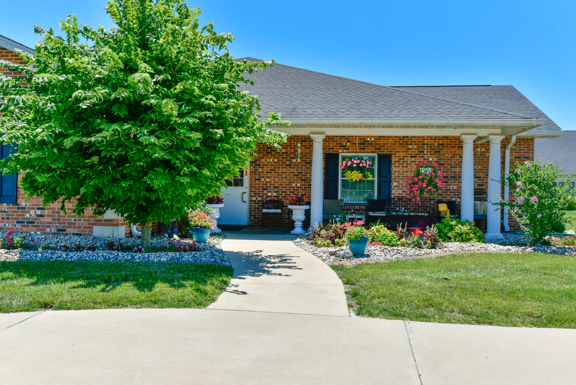 Front exterior view of a single-story brick building with a gray shingled roof, white columns supporting a small porch, and a concrete walkway leading to the entrance. The area is landscaped with green grass, a leafy tree, flower beds with colorful flowers, and potted plants under a clear blue sky.