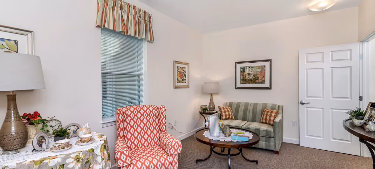 A cozy living room with a patterned red armchair, a green striped loveseat with two plaid pillows, a round wooden coffee table with decorative items, a side table with a lamp, and a window with blinds and a striped valance. The walls are white with framed artwork, and a white door is open to another room.