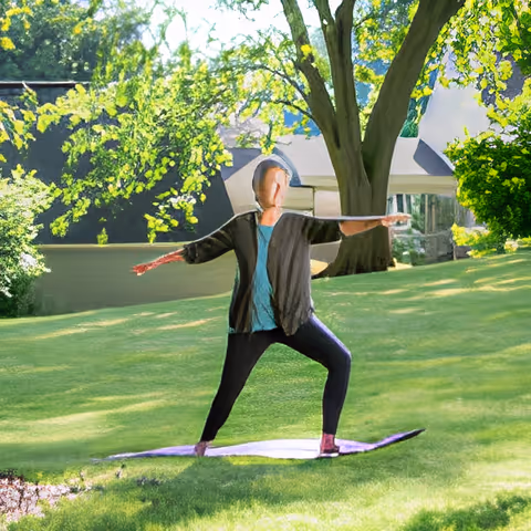A person practicing yoga outdoors on a purple mat on a grassy lawn surrounded by trees and greenery, with a building partially visible in the background.