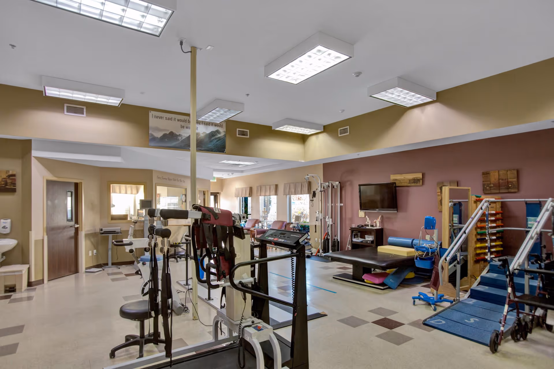 Bright rehabilitation gym in a senior facility with exercise machines, therapy tables, mats, and parallel bars.