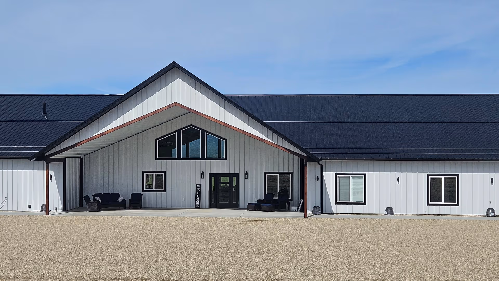 Front exterior of a white single-story building with a black gabled metal roof and a covered entrance with outdoor seating.