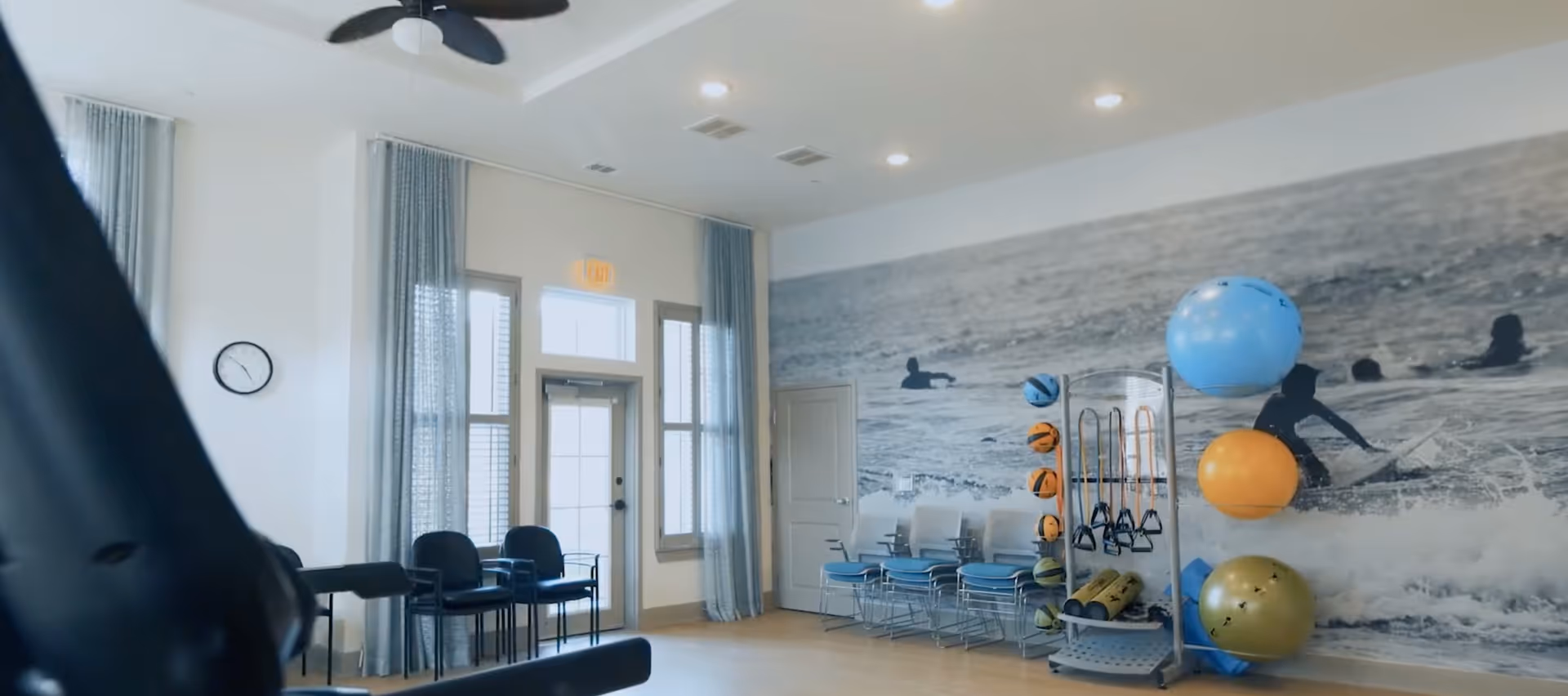 A bright exercise room with light-colored walls and floor, featuring a large wall mural of people surfing in the ocean. The room has exercise equipment including a rack with various sized exercise balls, resistance bands, and weights. There are several stacked chairs along the wall near a door with windows and blue curtains. A ceiling fan and a clock are also visible.