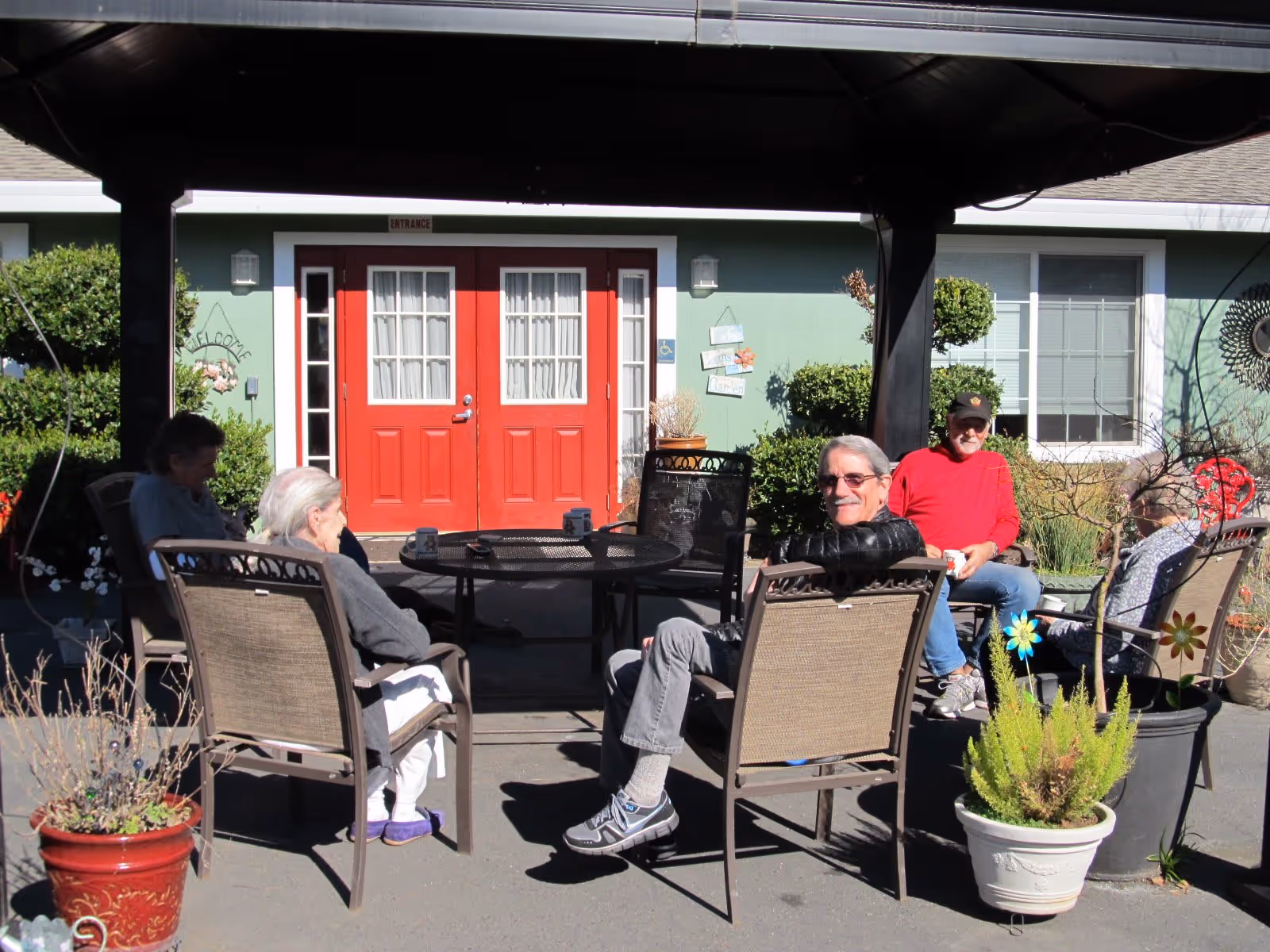 A group of elderly people sitting and chatting around a round outdoor table under a shaded structure in front of a building with red double doors and green walls. There are potted plants around the seating area and a welcome sign near the entrance.