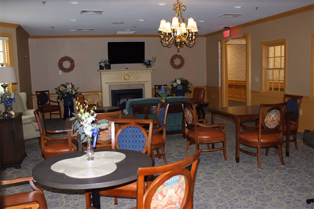 A cozy common area in an assisted living facility featuring multiple wooden tables and chairs with patterned cushions, a fireplace with a TV mounted above it, floral arrangements on tables and around the room, a chandelier hanging from the ceiling, and soft lighting creating a warm atmosphere.