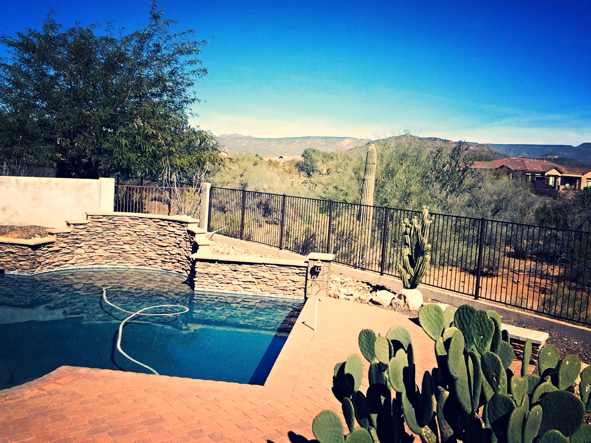 Outdoor view of a swimming pool with a stone wall feature, surrounded by a brick patio. In the background, there is a black metal fence, desert vegetation including cacti, and distant mountains under a clear blue sky.
