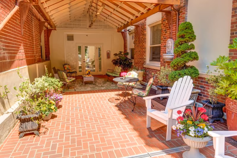 Covered brick patio courtyard with potted plants, Adirondack and metal chairs, and small tables alongside a brick building.