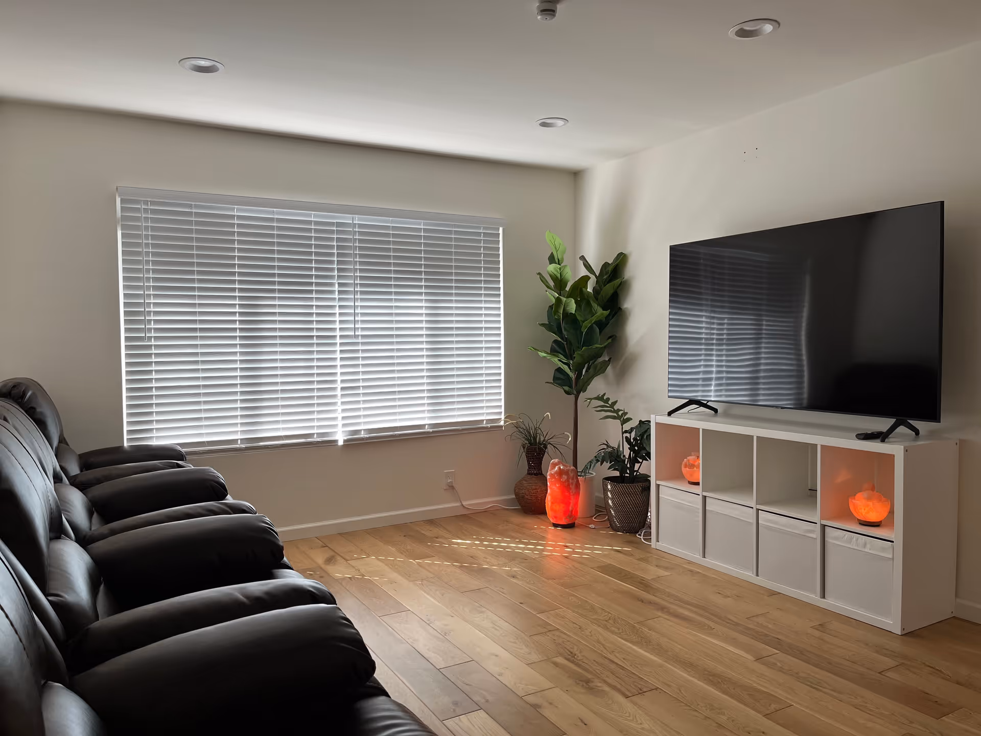 A living room with a row of black leather recliner chairs facing a large flat-screen TV on a white storage unit. The room has wooden flooring, a large window with white blinds, and several potted plants including a tall leafy plant and smaller ones. Two glowing orange salt lamps are placed on the storage unit and the floor.