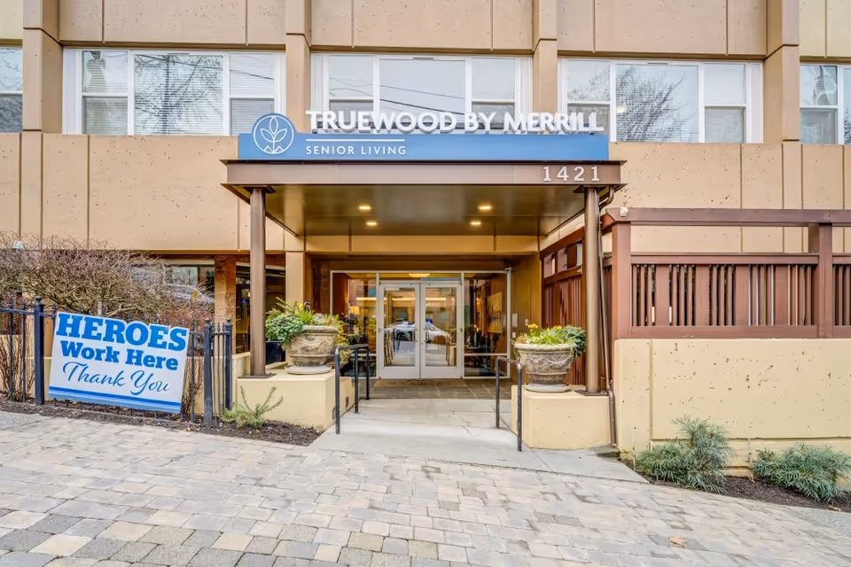 Entrance to the Truewood by Merrill senior living building with a canopy, address 1421, and a 'HEROES Work Here Thank You' sign.