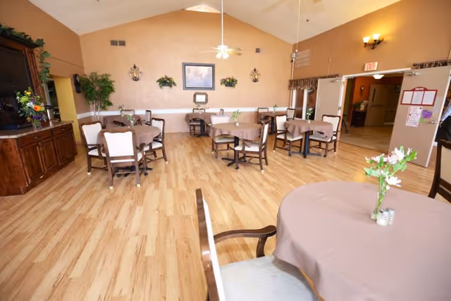 A spacious dining room with several round tables covered with beige tablecloths, each table decorated with a small vase of flowers. The room has wooden flooring, beige walls, wall-mounted light fixtures, and a ceiling fan. There is a wooden cabinet with a TV on the left side and an open doorway leading to another room on the right.