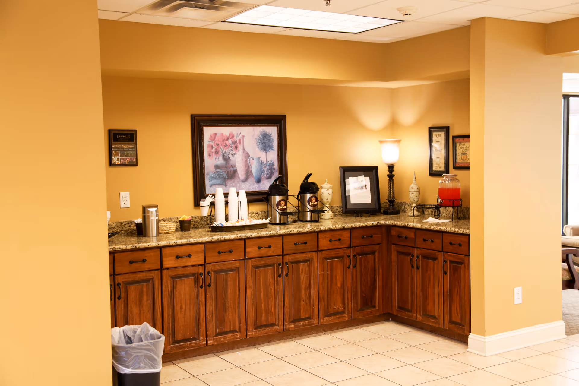 A corner kitchenette area with wooden cabinets and granite countertops. On the counter, there are two coffee dispensers, disposable cups, a lamp, framed pictures, decorative vases, and a beverage dispenser with red liquid. The walls are painted beige, and the floor is tiled. A trash can is visible on the left side.