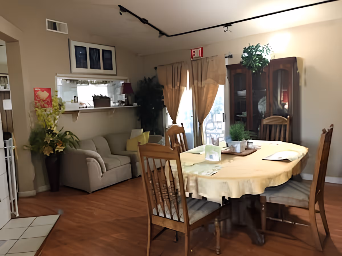 Dining room with a round table covered by a yellow tablecloth surrounded by wooden chairs, a china cabinet, and a small sofa near a pass-through window.