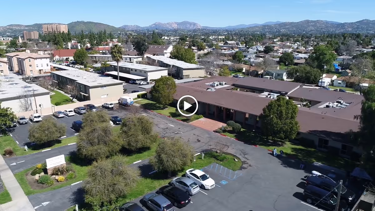 Aerial view of Magnolia Post Acute Care facility showing multiple buildings with brown roofs, parking lots with cars, landscaped areas with trees and grass, and a surrounding residential neighborhood with mountains in the background under a clear sky.