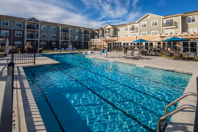 Outdoor swimming pool with clear blue water at a senior living facility, surrounded by lounge chairs, tables with umbrellas, and a three-story building in the background under a partly cloudy sky.