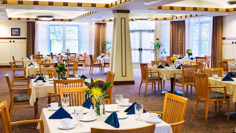 A bright and spacious dining room with multiple round tables covered with white tablecloths, each set with glassware, white cups, plates, silverware, and navy blue folded napkins. Fresh flower arrangements decorate the tables. The room has large windows with brown curtains allowing natural light to fill the space, and wooden chairs with patterned cushions surround the tables.