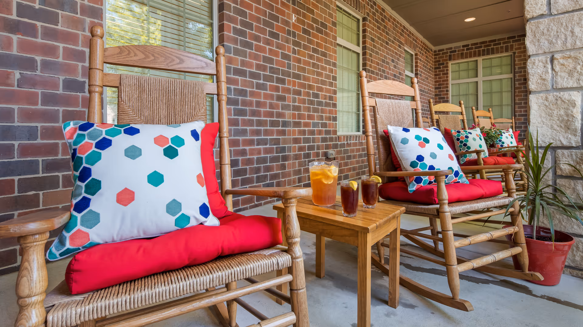 A row of wooden rocking chairs with red cushions and colorful hexagon-patterned pillows on a covered porch with brick walls and windows. A small wooden table holds a pitcher and glasses of iced tea with lemon slices. A potted plant is placed near the chairs.