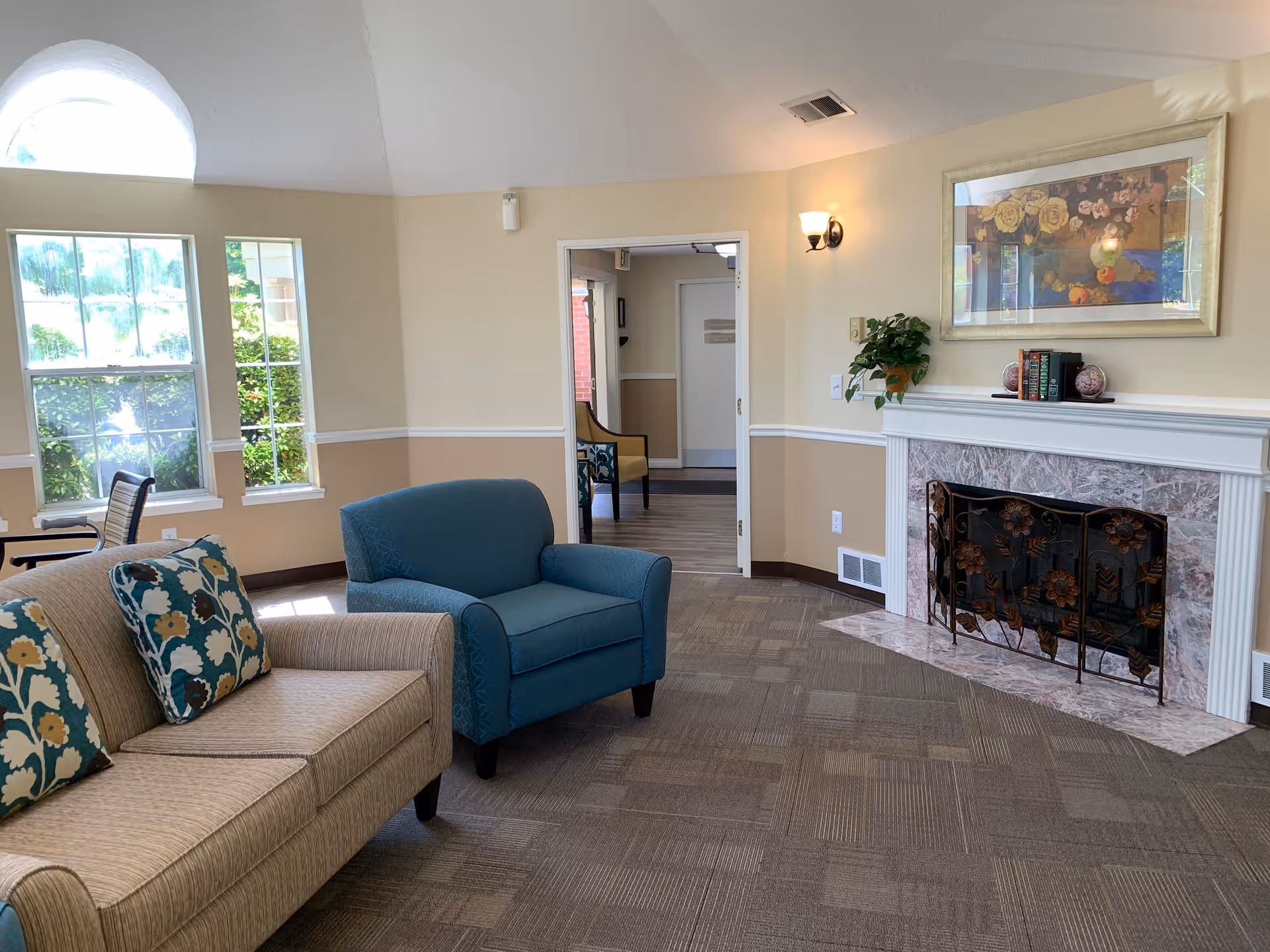 A cozy living room area in a senior living facility with a beige sofa adorned with floral patterned pillows, a blue armchair, a fireplace with a decorative screen, a framed floral painting above the mantel, and a small plant. Large windows allow natural light to brighten the room.