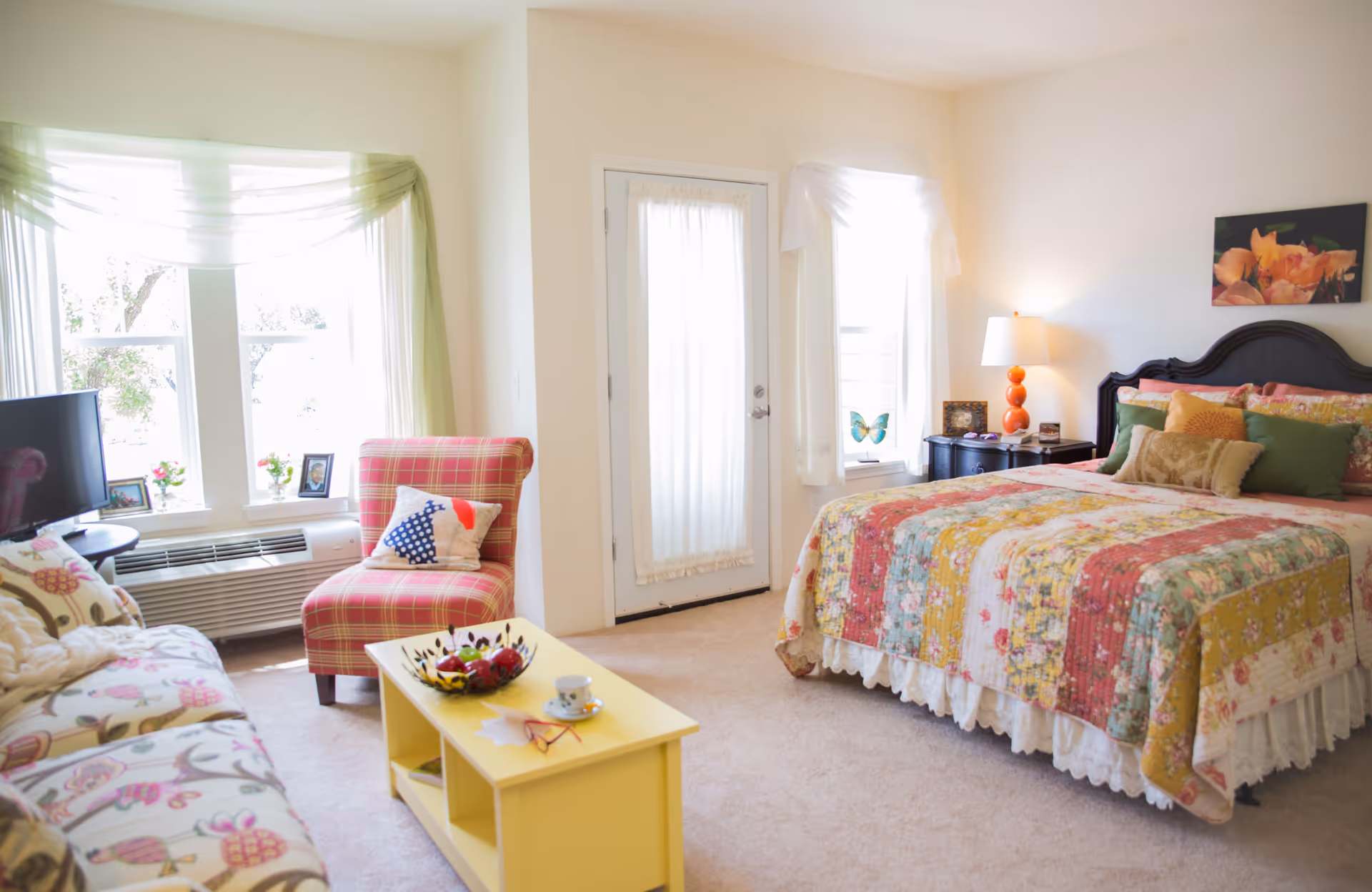 A bright and cozy bedroom with a colorful patchwork quilt on the bed, a nightstand with a lamp, a floral sofa, a red plaid armchair with a decorative pillow, a yellow coffee table with a bowl of fruit and a teacup, and large windows with sheer curtains letting in natural light.