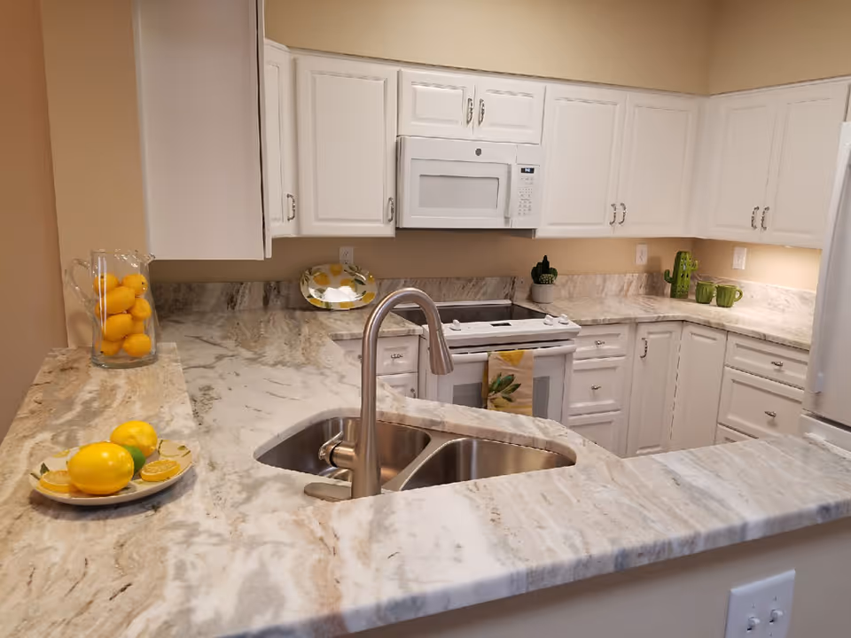 Modern kitchen with white cabinets, marble countertops, a double stainless steel sink with a curved faucet, a white microwave above a white stove, and decorative items including a plate and pitcher with lemons and green mugs on the counter.