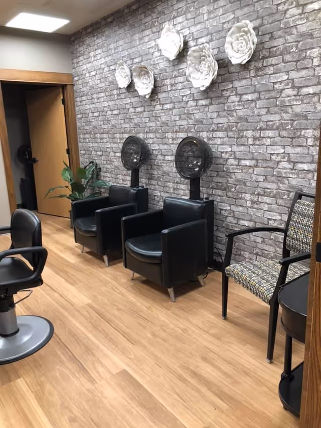 A salon-style interior with two black hooded hair dryers and chairs against a faux brick accent wall decorated with white flower sculptures.